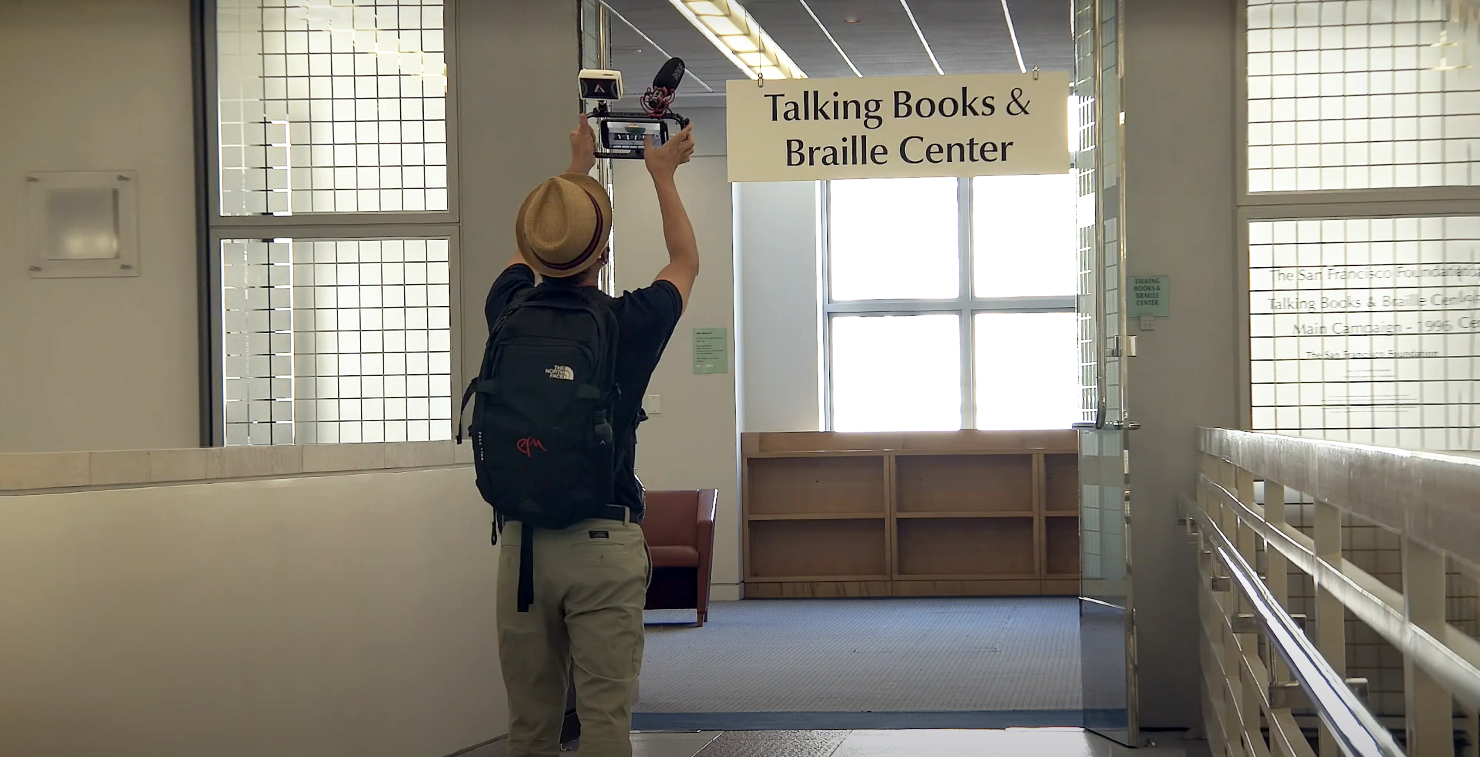 Image of a man wearing a hat and holding a film camera above head as he walks into the Talking Books and Braille Center at the SFPL