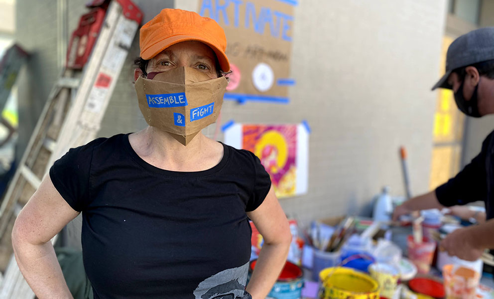 Image of artist wearing an orange ball cap and a face mask made out of brown paper and reads Assemble & Fight