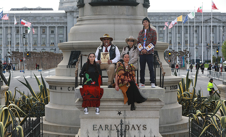 Five indigenous individuals standing and sitting on an empty plinth