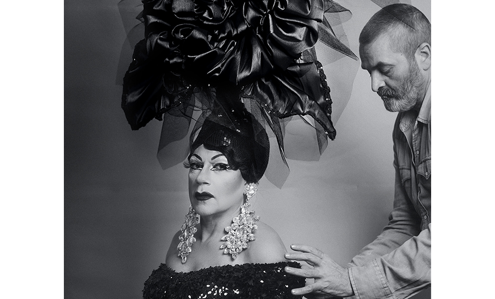 Black and white photograph of Juanita MORE! in a black off the shoulder dress and wearing an extravagantly tall black feather headdress. She is seated in three-quarter view of the camera, Mr. David, seen in profile, is slightly off frame, standing and adjusting Juanita's gown.