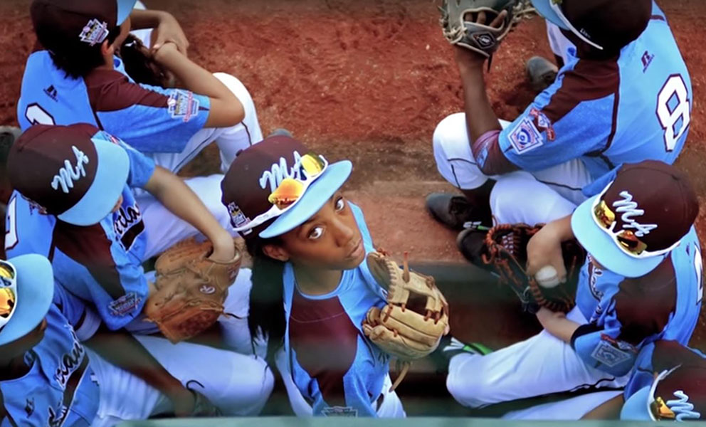 image from above of a baseball team in blue uniforms. The central figure, a young woman, is looking up directly into the camera.