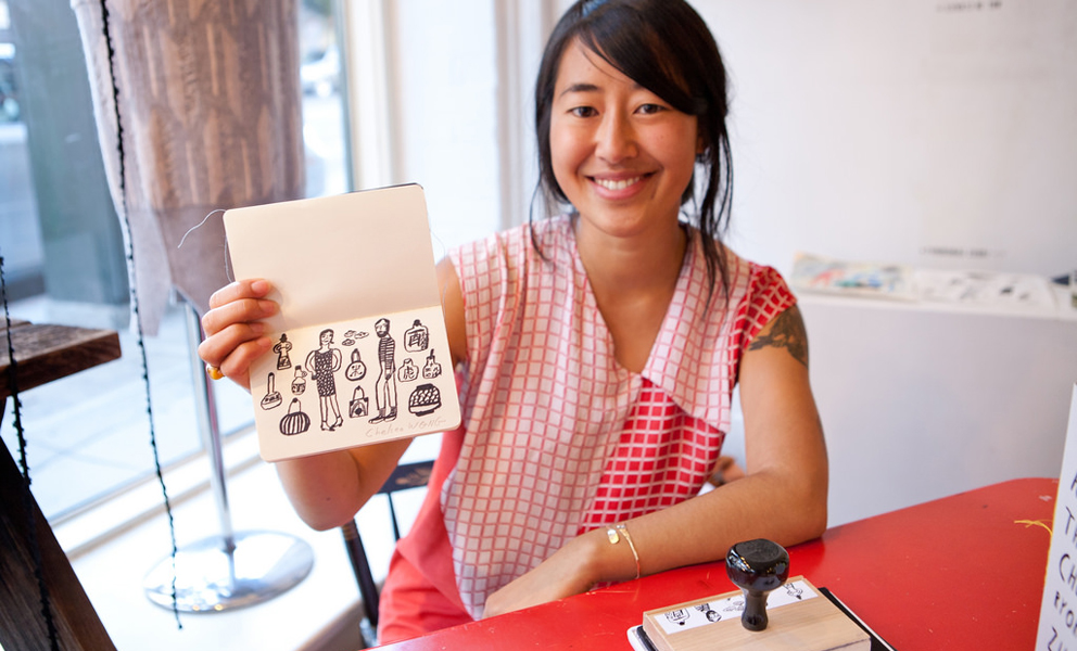 Image of smiling woman wearing red holding a small book with artist designed stamp in it. 