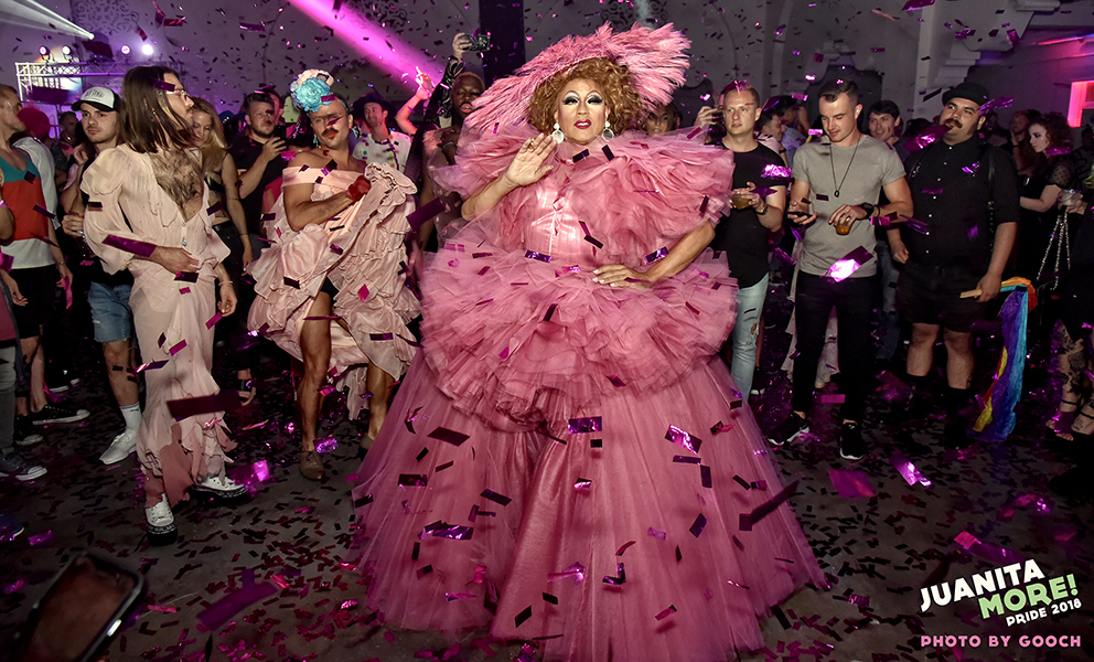 Juanita MORE!, in a voluminous pink ruffled dress and a matching hat, stands in the center of a crowd indoors with purple confetti flying in the air.