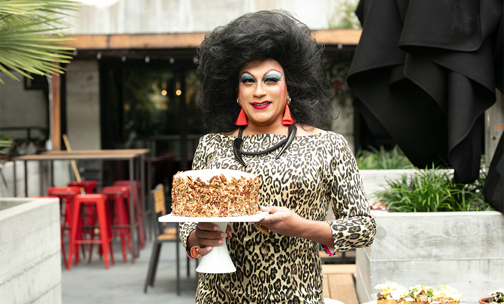 Juanita MORE! stands holding a decorated cake on a white cake stand, smiling. She wears a long-sleeved leopard print dress with a chunky black necklace. She is standing in an outdoor patio.
