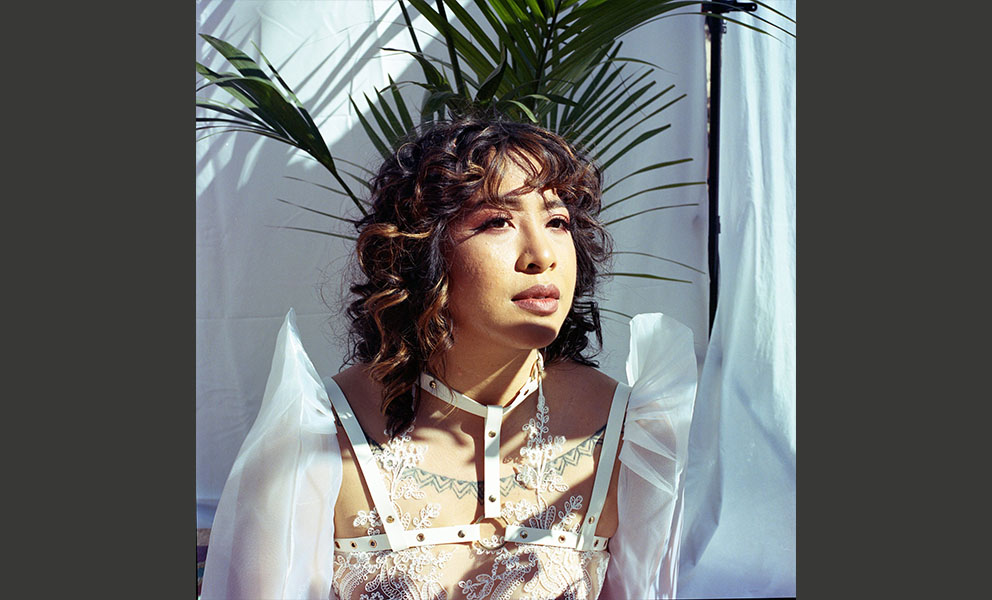 Image of the artist wearing a white mesh shirt with floral pattern and sculpted shoulders seated in front of palm tree.