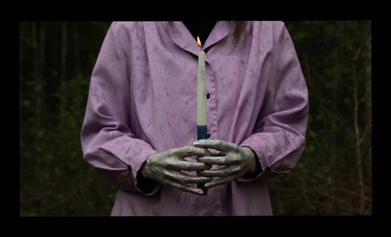 image of person wearing lavender colored shirt, holding a candle with painted silver hands. 