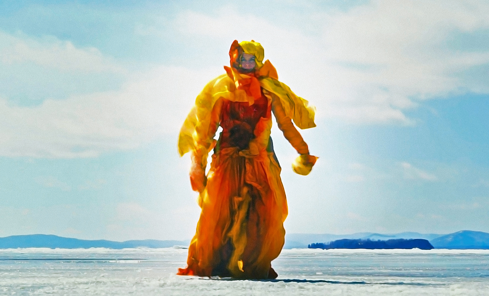 A figure covered except for the face in yellow and orange fabric approaches the camera, walking on a frozen lake