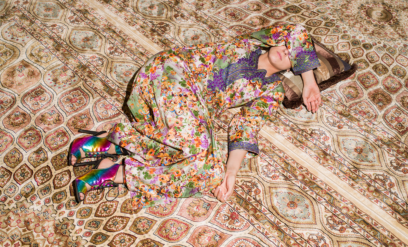 A woman in a floral print lays across a patterned rug, covering her face. 