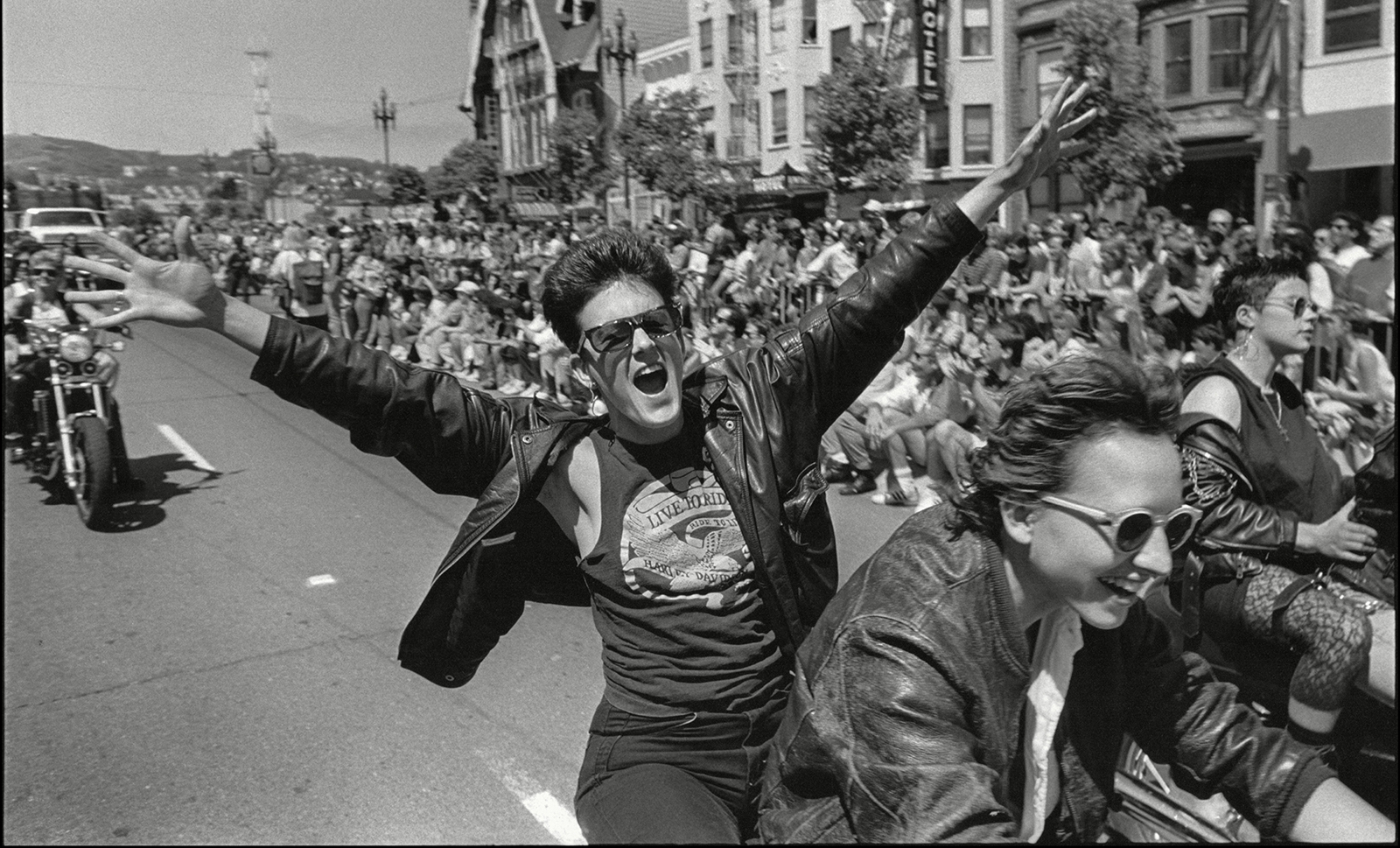 A black and white image of a woman on the back of a motorcycle with her arms raised up.