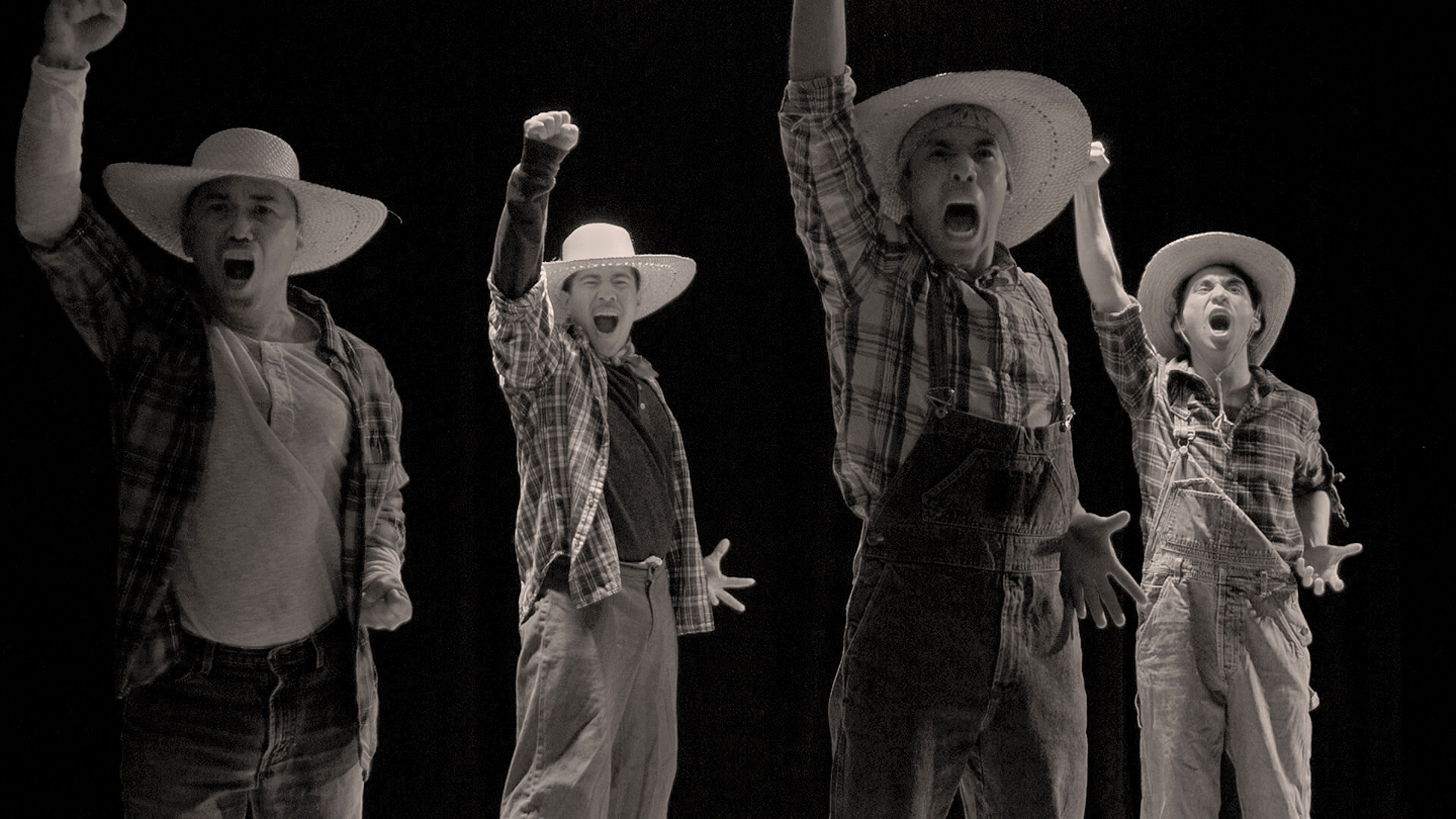 A black and white photo of four men dressed in laborers clothing with their fists raised up in the air and with mouths open shouting.