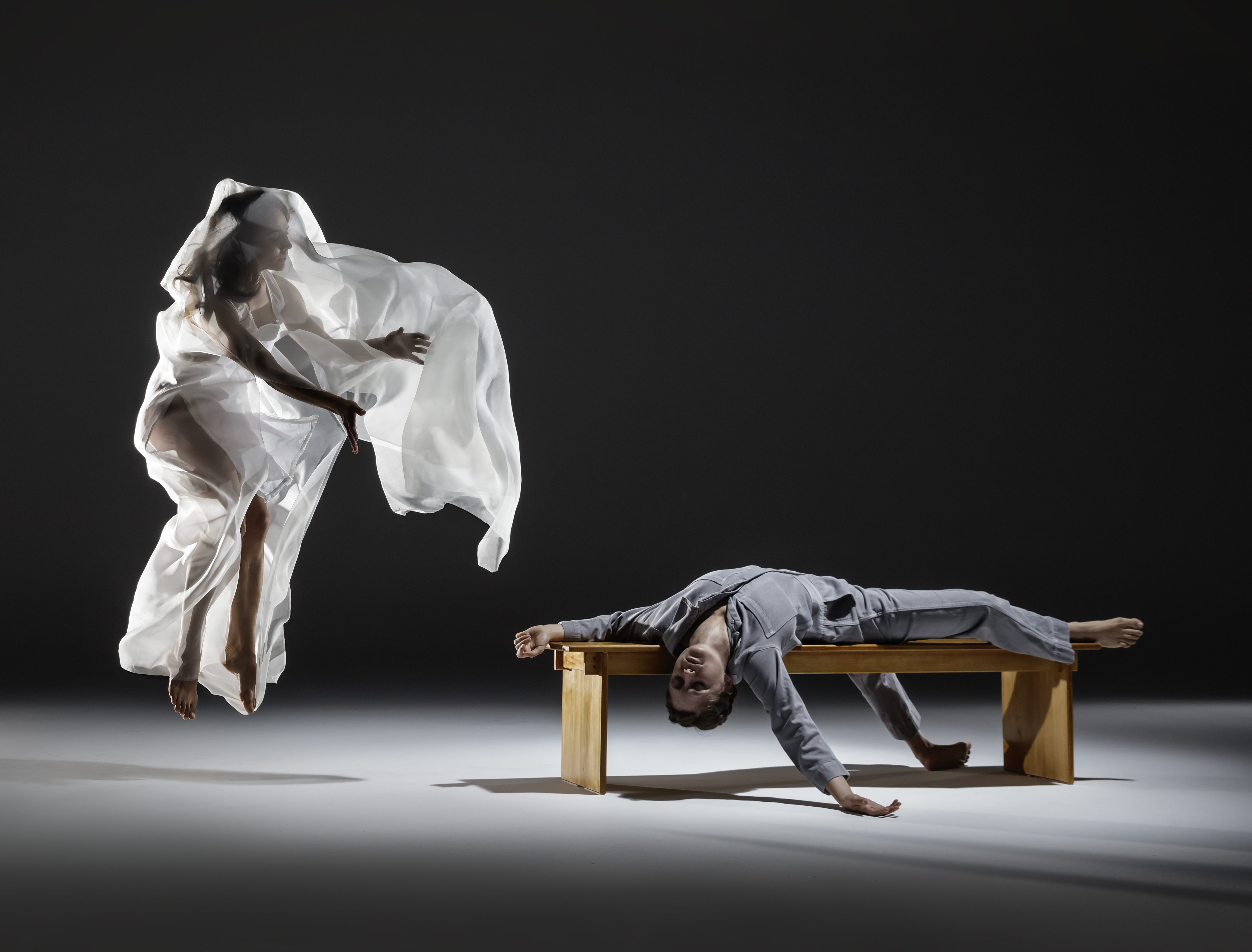 Two performers on a dark stage, one covered in a white sheet jumping in the air and one in grey jumpsuit laying on a wooden bench