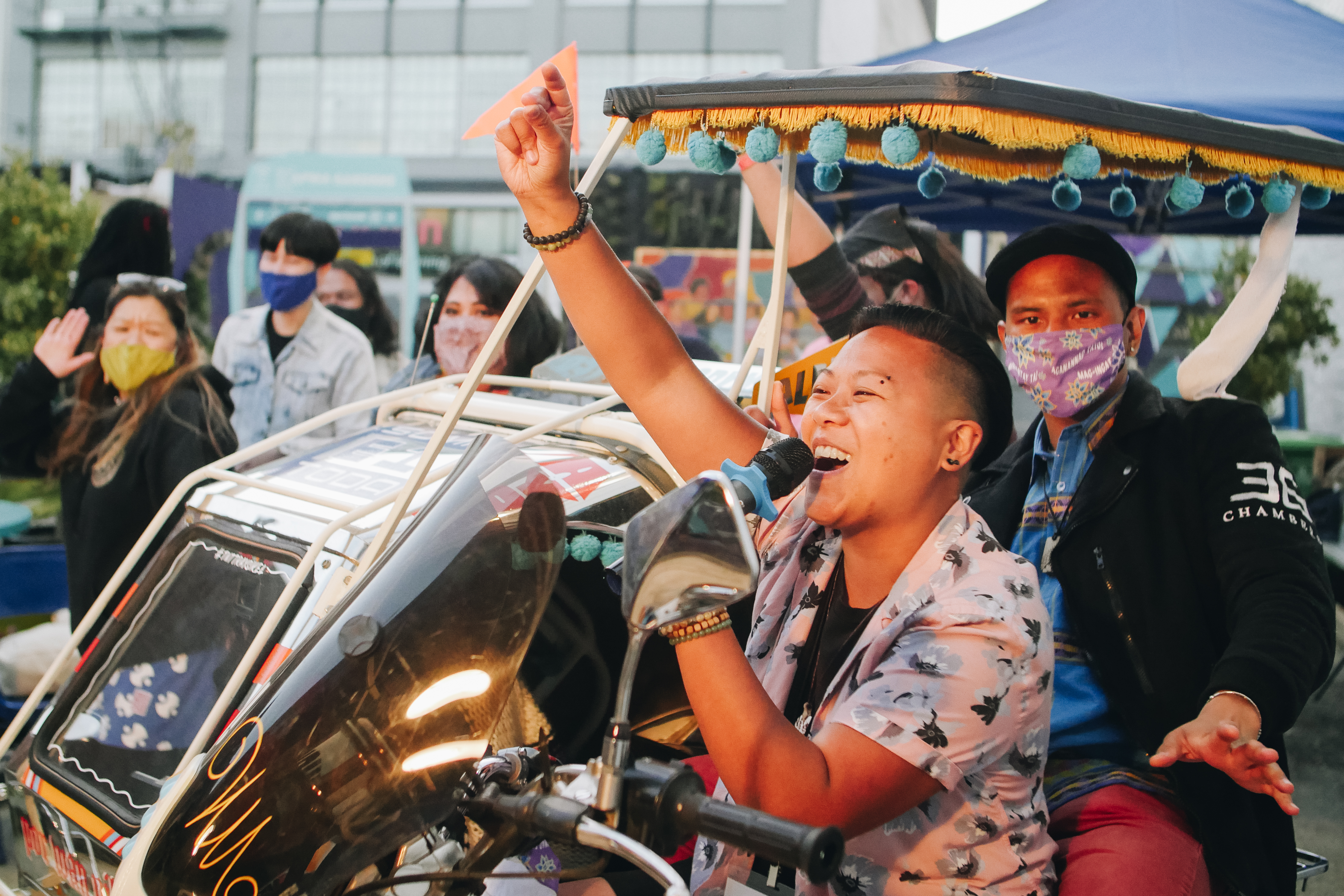 In the foreground a Philipinx person with a side shaved haircut and facial piercings holds a microphone in their left hand and raises their right in a snap. Behind them, two other people sit in a decorative tricycle, a croud cheers around them in the background.