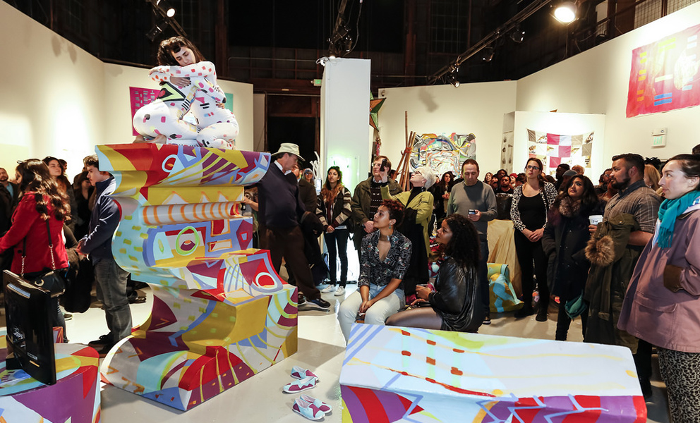 Visions Into Infinite Archive opening event at SOMArts A performance artists crouches on top of a colorful geometric sculpture in the middle of a crows of onlookers
