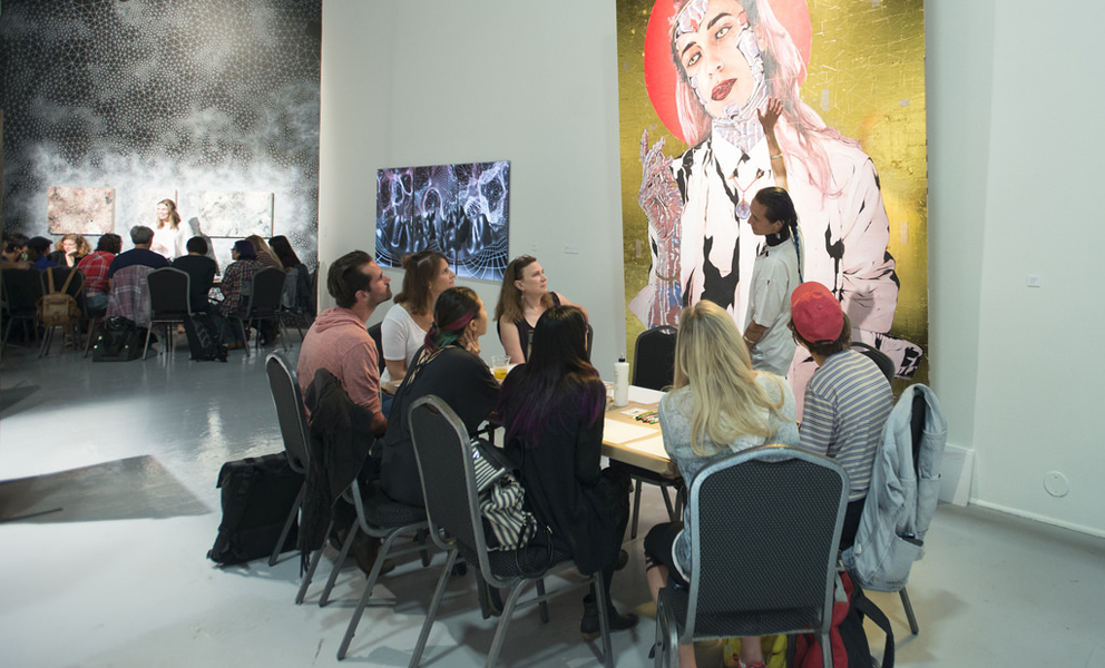 Group of people gathered around a table listening to a speaker in front of a large figurative painting. 