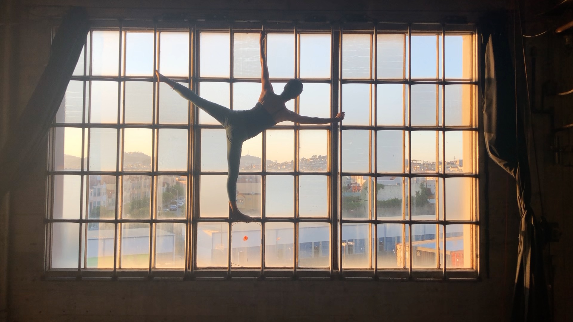 Joanna Haigood in Zaccho Dance Theatre's film RECORD TWENTY TWENTY for the 2020 San Francisco Aerial Arts Festival. Color photograph of a multi-paned glass window with a person's silhouette in the middle of the frame who is standing on the right leg and outstretched in a posed position.