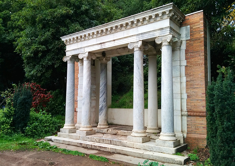 A sculpture that mimics a classical building facade with columns, surrounded by trees and hedges, seen from a slight angle.