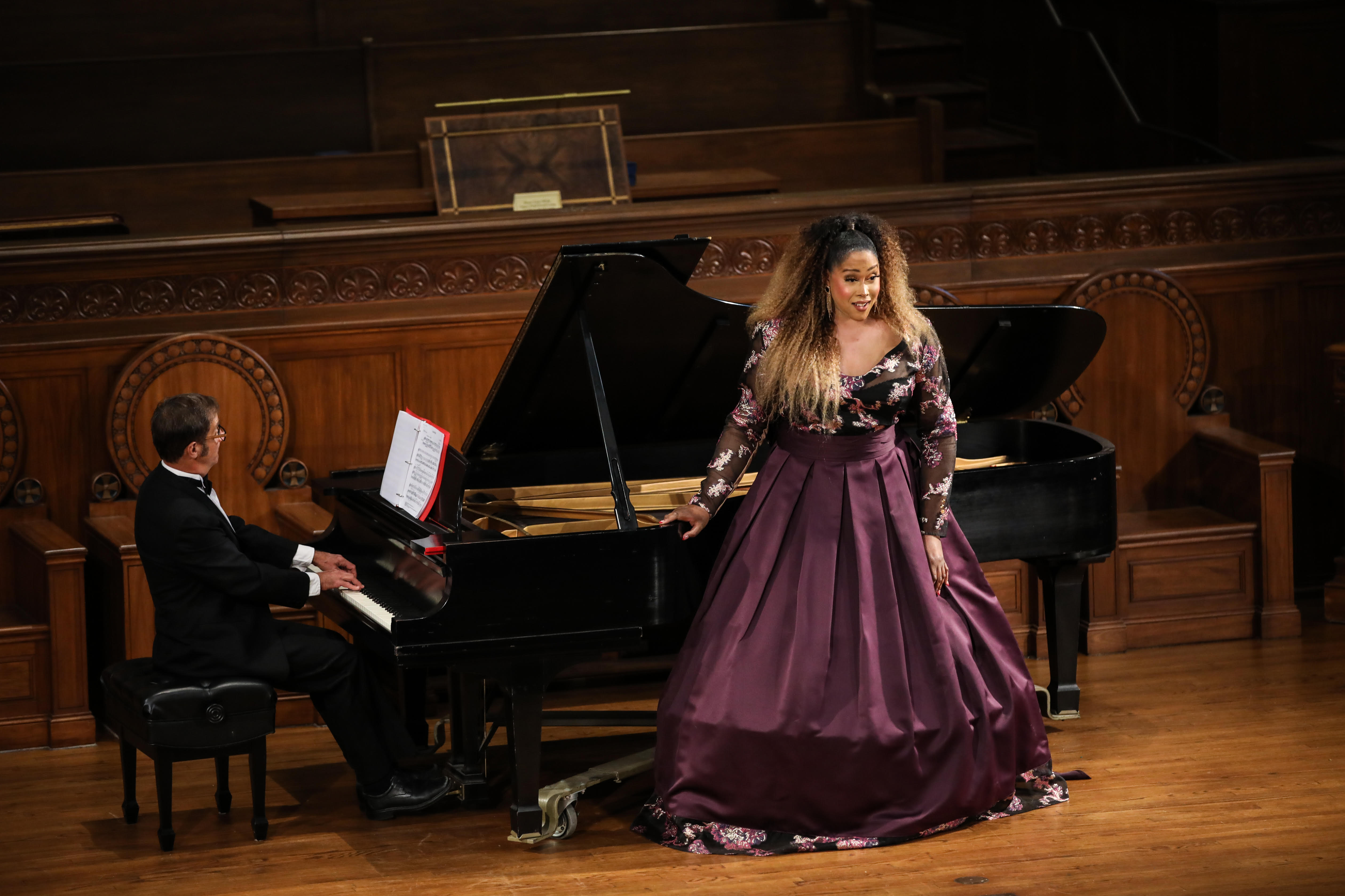 Breanna Sinclaire | Photo Credit Misti Layne Black woman opera singer with long blond and black hair in a floor length long-sleeved hooped burgundy dress standing in front of a grand piano on stage