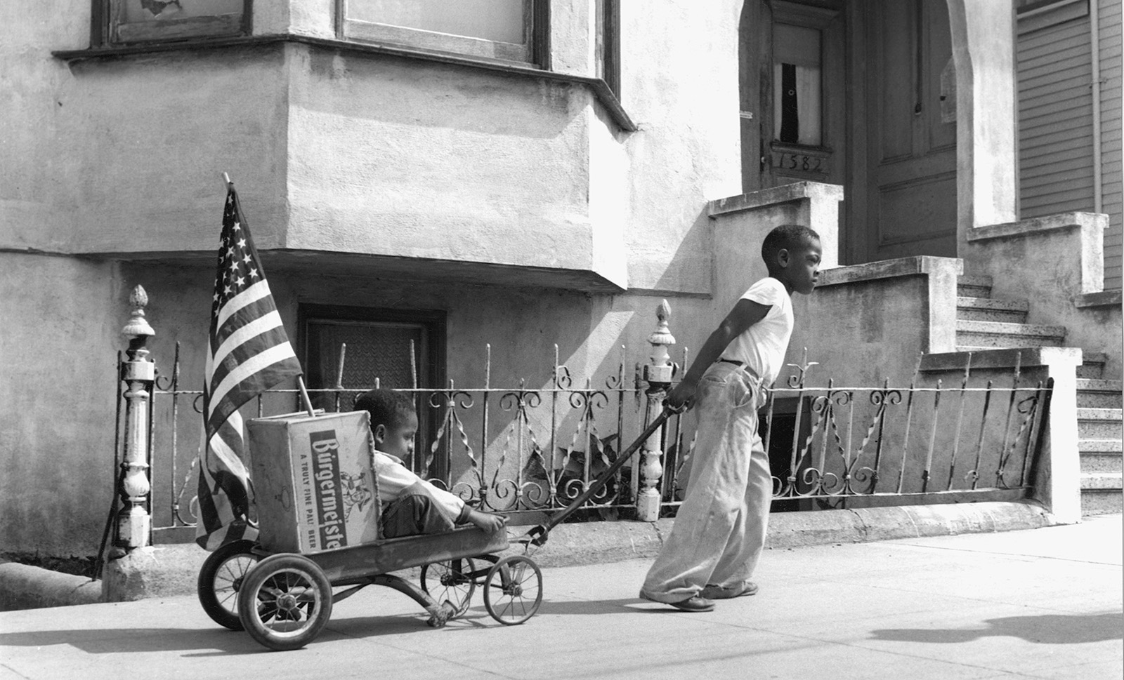 A black and white photograph of a young Black boy pulling on a wagon, walking towards the right of the fame. On the wagon, sits another Black boy and a box with an American flag coming out of it. The boys are in front of a residential building in Hunter's Point. 