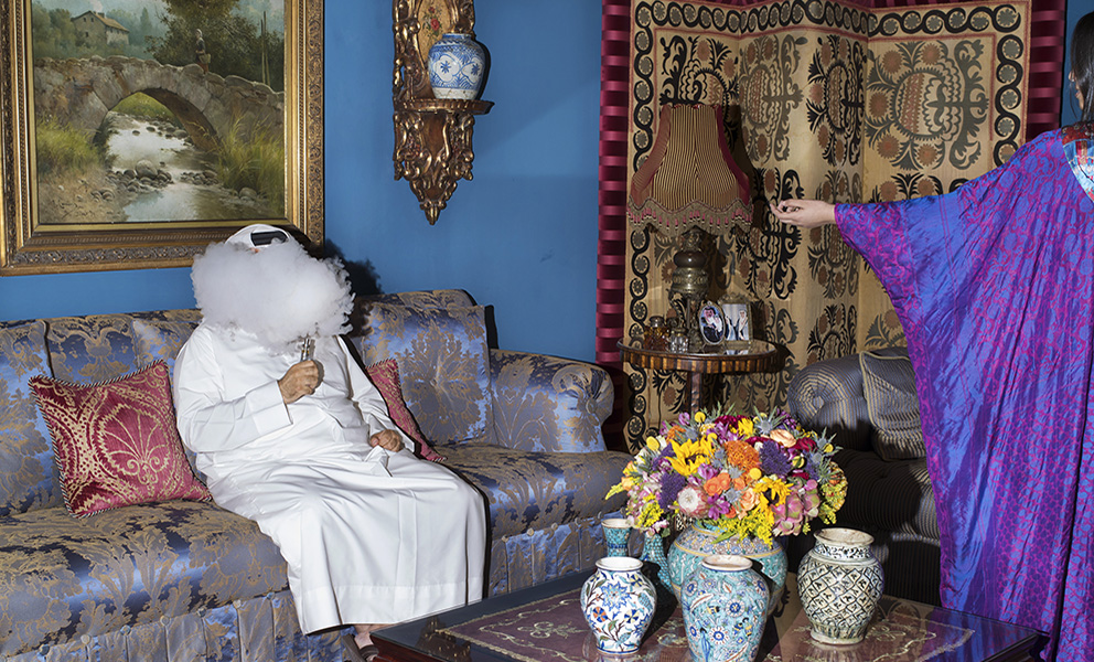 Person sitting on couch in an elaborately decorated living room smoking. Woman in purple gown gesturing toward person on the couch.