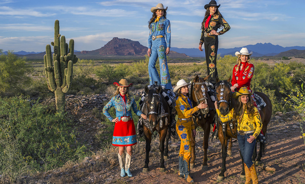 Image of six women dressed in elaborate cowgirl outfits with horses in the dessert