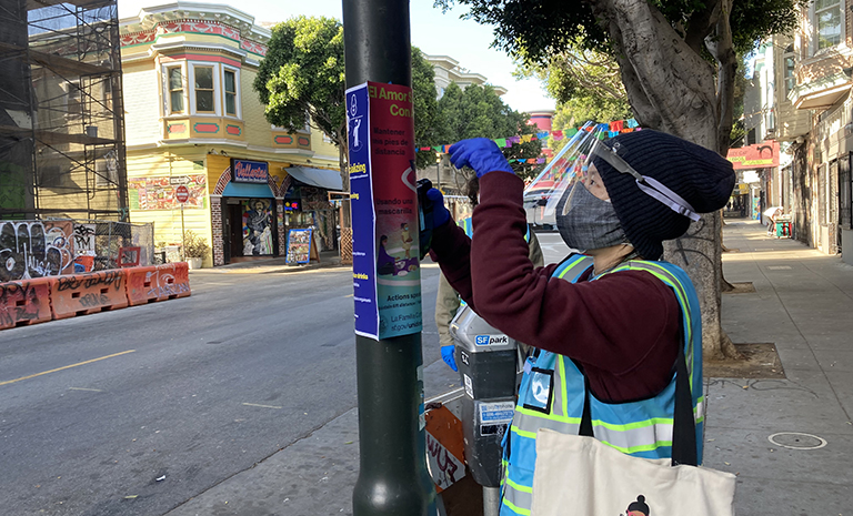 An Asian person wearing a face mask, face shield, black hat and blue safety vest is taping up infographic signs on a lightpole on a street.