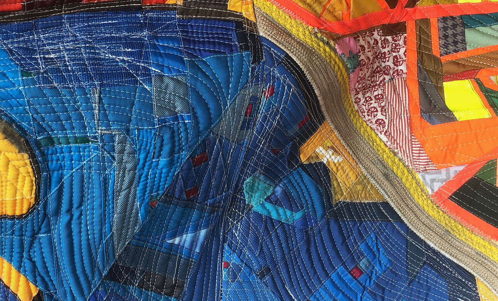 A close up of a richly textured quilted textile work, largely made of blue, orange and yellow scavenged fabrics. 