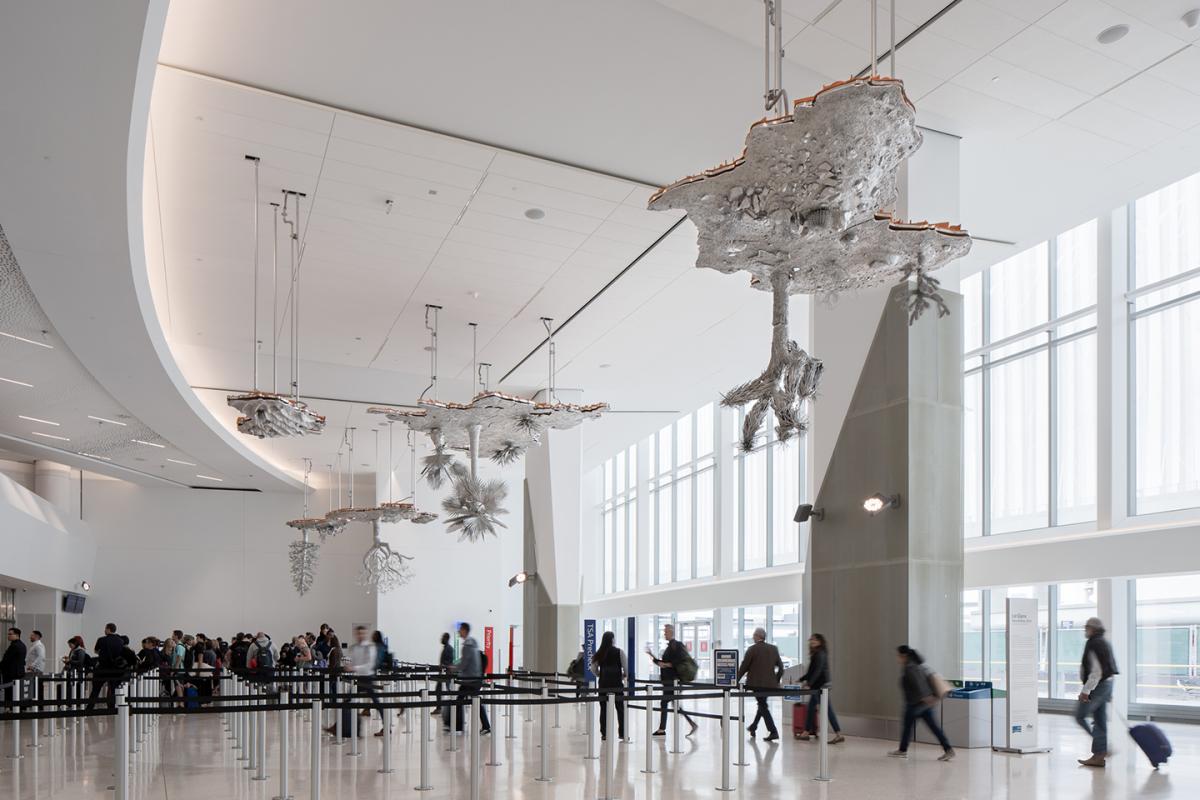 A view of a suspended metal sculpture by Liz Glynn. The sculpture, hanging from the ceiling of SFO resembles a landscape with trees and brush, looking like an inverted landscape.