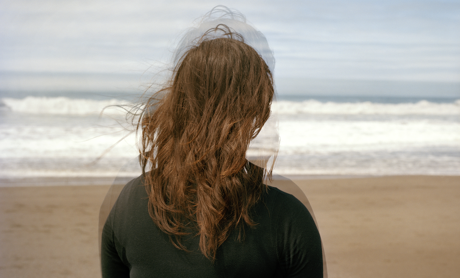 An image of a woman, seen from behind, with long brown hair standing at the beach. The figure is facing the ocean. The image is composited with another image of the woman overlaid, and slightly askew so that the image has a blurred effect.