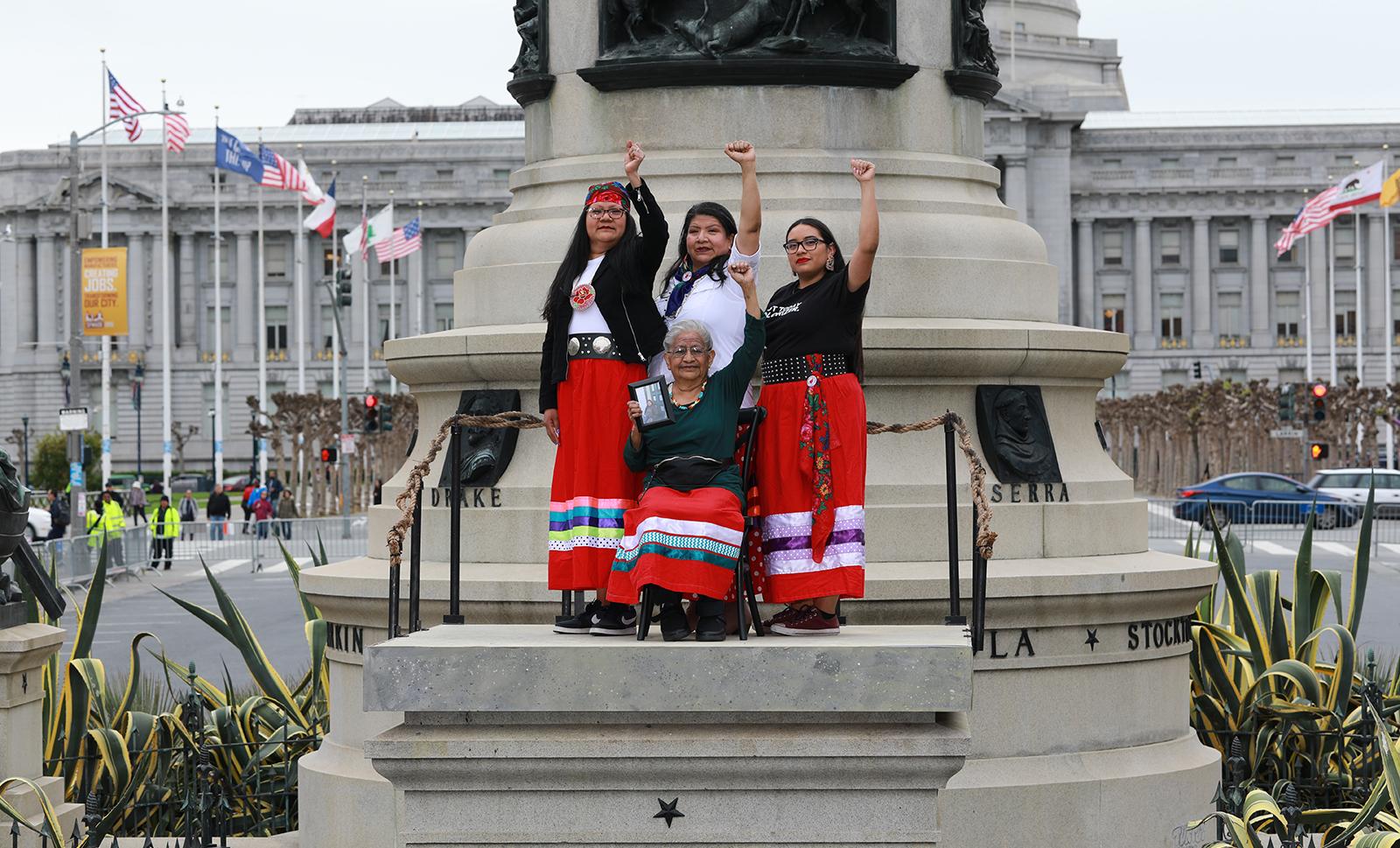 Four Indigenous women standing on an empty pedestal wearing traditional regalia 
