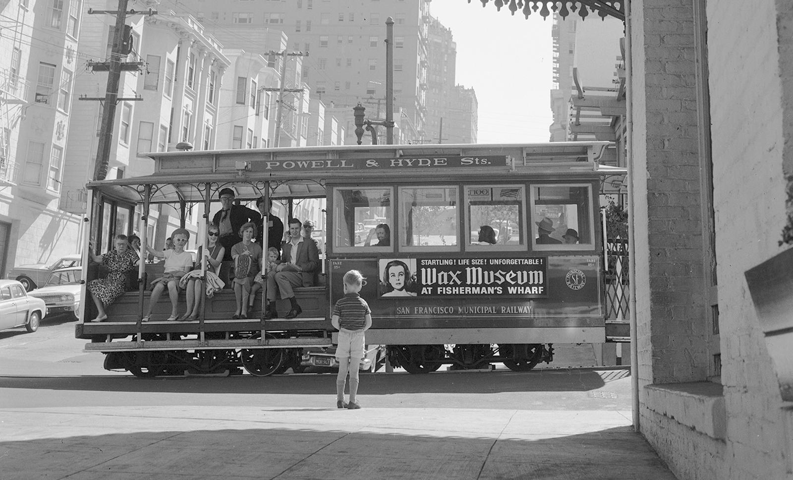 From an image from 1965: a cable car is stopped, and beside it is a young boy with their back to the viewer, looking at the cable car.