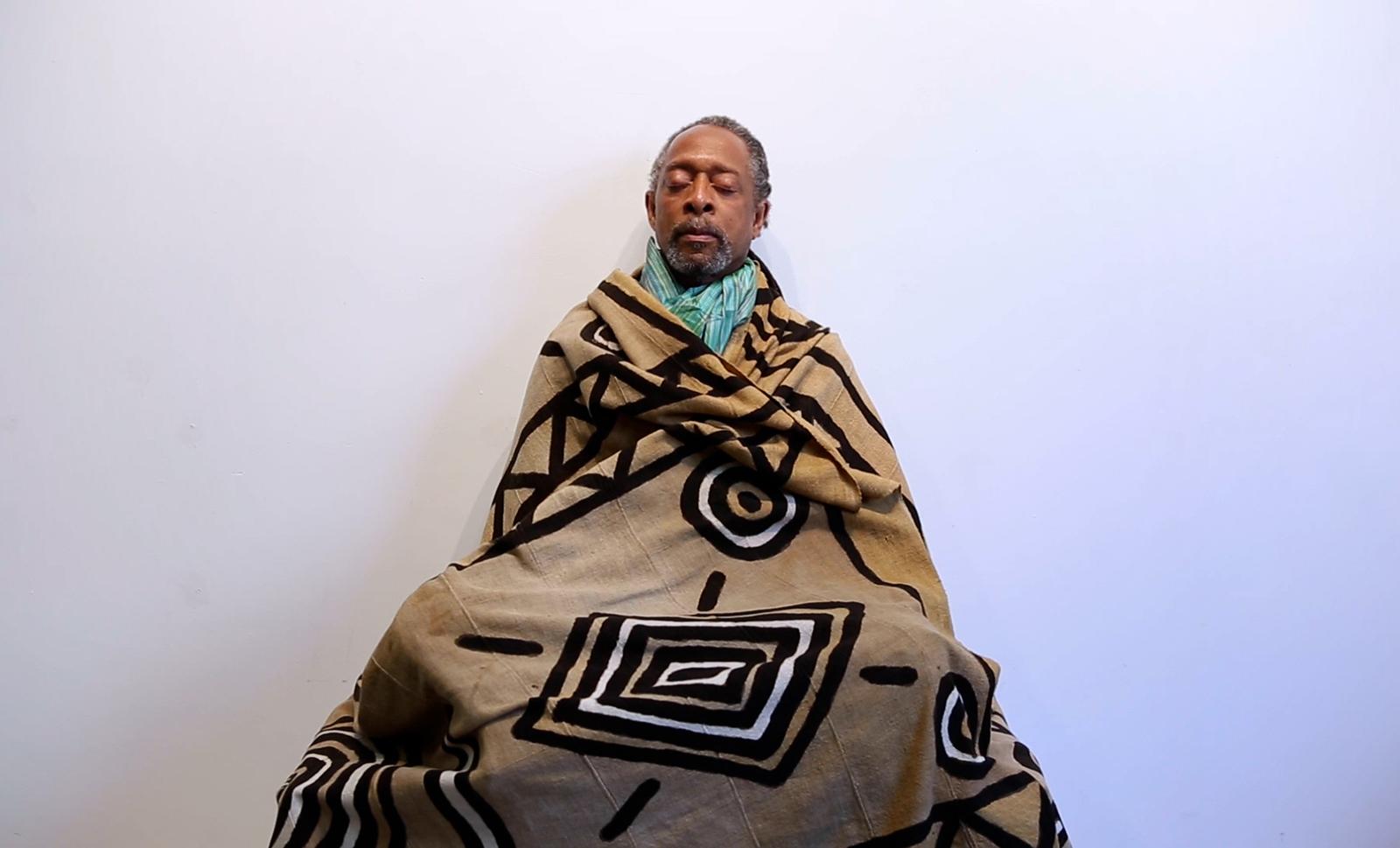 A Black man sitting, wrapped in tan blanket with black and white patterns, wearing a light blue scarf, and with his eyes closed in meditation.